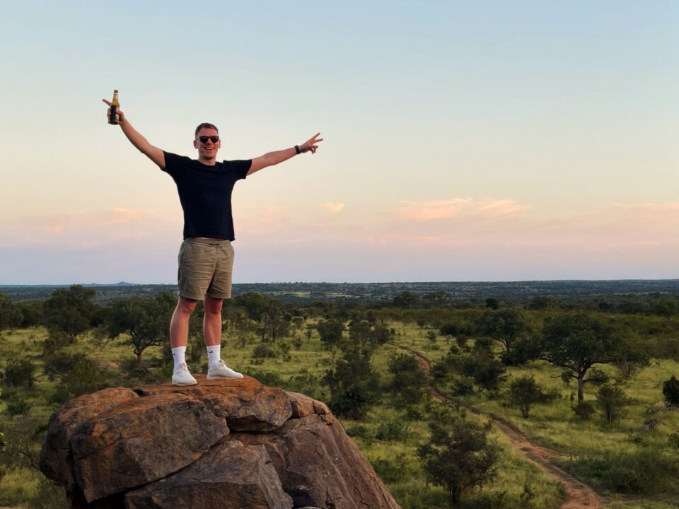 A man smiles at the camera standing on a large boulder above the bushveld. It's dusk and the sky is a little pink.