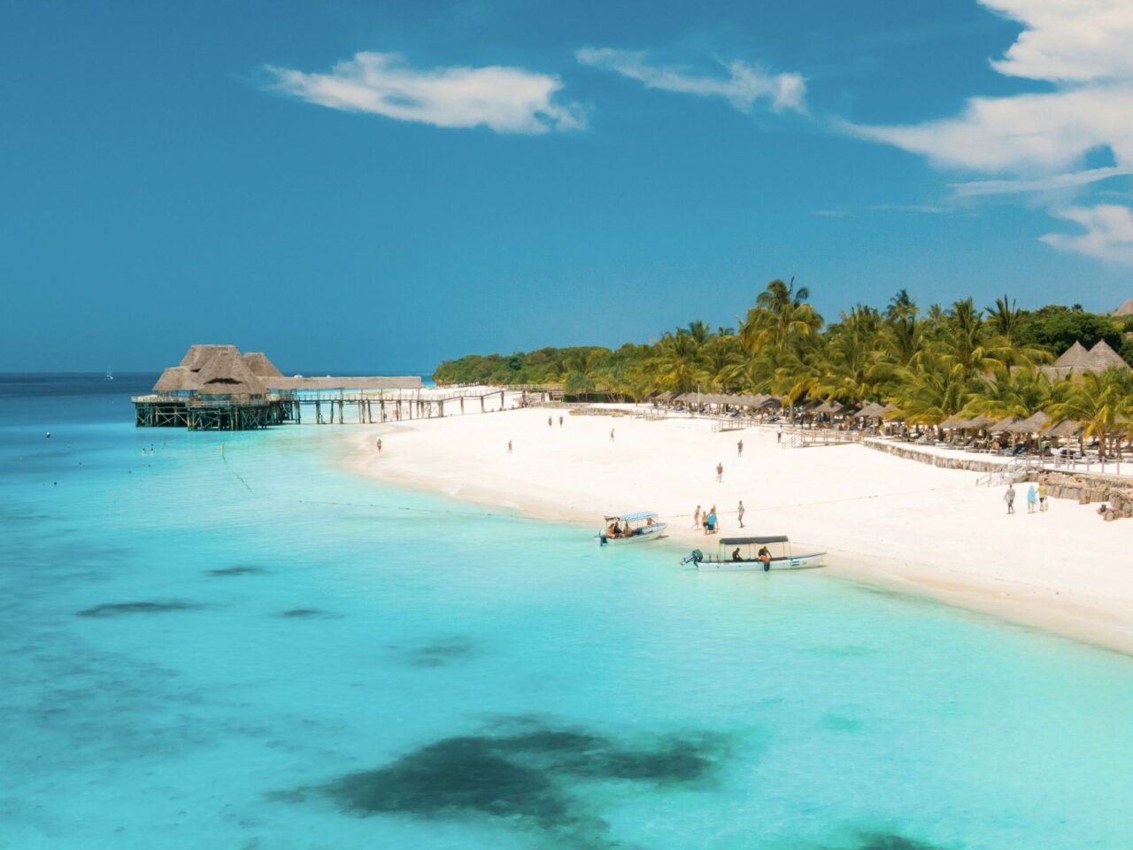 A brilliant white sand beach is lapped by clear blue waters. People are playing in the water on a boat and the sky above is blue. This is a beach in Zanzibar, Tanzania.