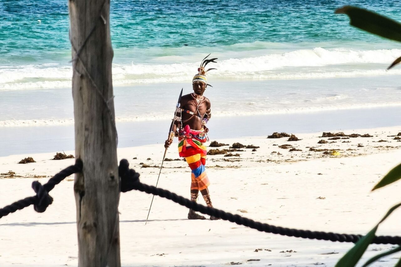 A man stands on a white sand beach in colourful tribal dress. He holds a spear and has feathers on his head. Behind him is the blue sea. This is Diani Beach in Kenya.