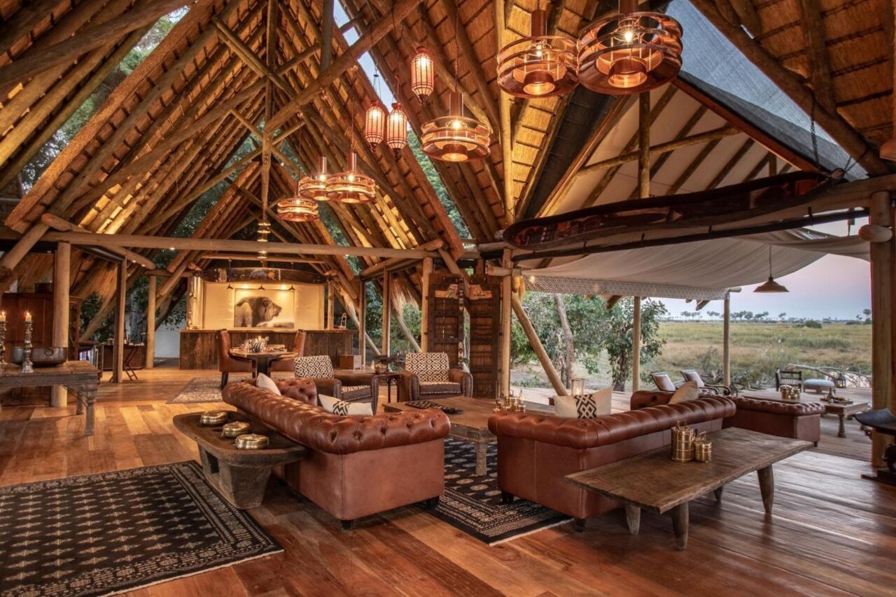 Open-air luxury lodge interior in Botswana featuring a thatched A-frame roof, leather couches, rustic wood furniture, and warm ambient lighting. The lodge overlooks a grassy savanna through wide openings, blending indoor comfort with the surrounding wilderness.