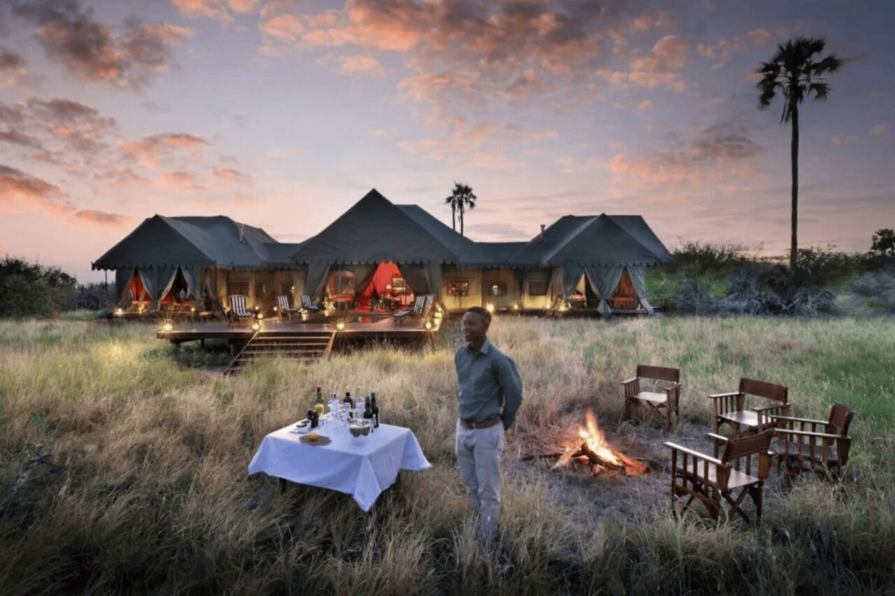 Luxury safari tented camp at sunset, with canvas-roofed pavilions lit warmly from within and surrounded by tall grass. A fire pit with wooden chairs and a table set for drinks are in the foreground, where a staff member stands smiling near the glowing campfire. The luxury camps are one of our top five reasons to visit Botswana.