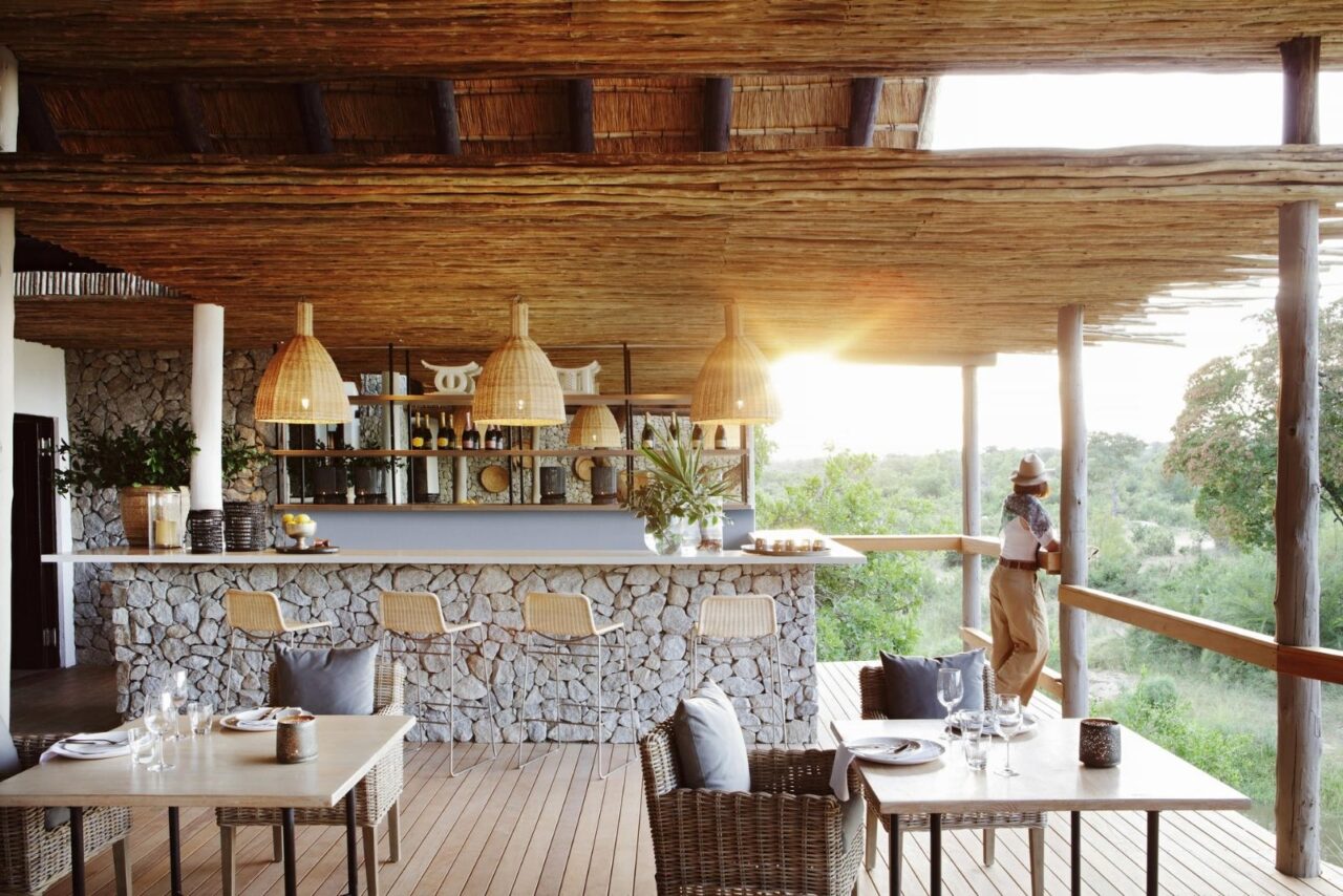 A woman stands on a semi open air terrace beside a bar looking down at the watering hole below. There are tables and chairs. The bar and space are stone and wood.