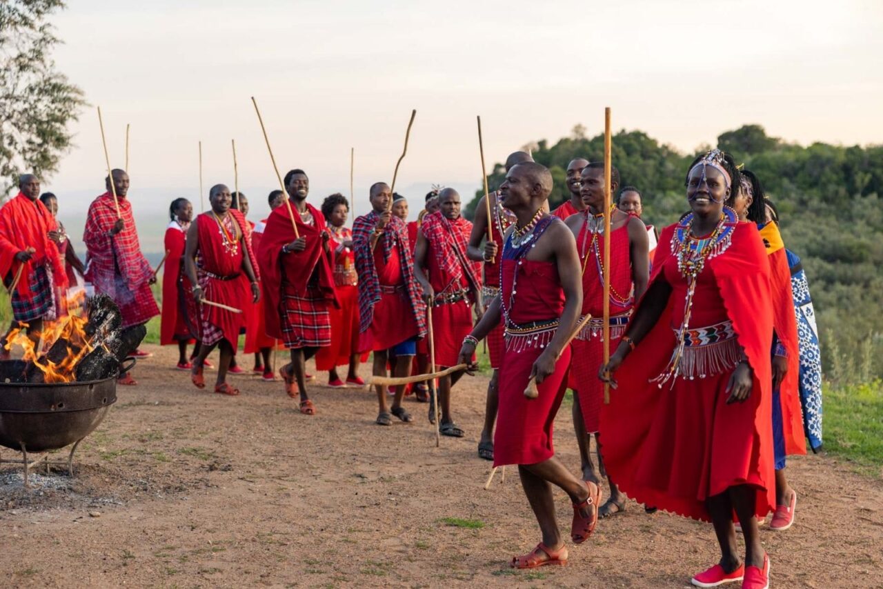 A group of Maasai men and women dressed in vibrant red shukas and traditional beadwork walk in a procession near a crackling outdoor fire, holding wooden staffs and smiling as they celebrate. The backdrop shows green vegetation under a soft evening sky, capturing the cultural richness of the Maasai community.