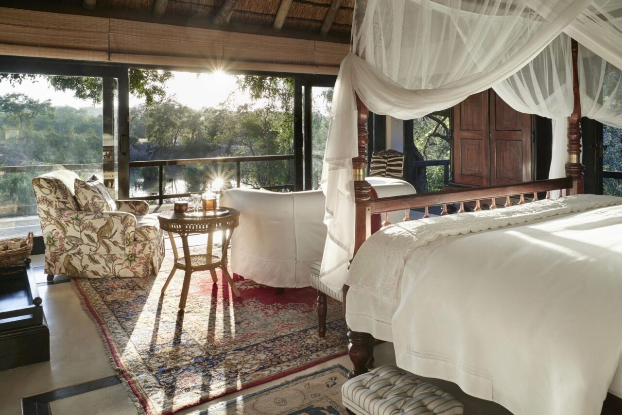 Interior of a luxury suite at Royal Malewane with a white canopied bed, red-patterned rug and armchairs. Sunlight streams through the large window illuminating the terrace.