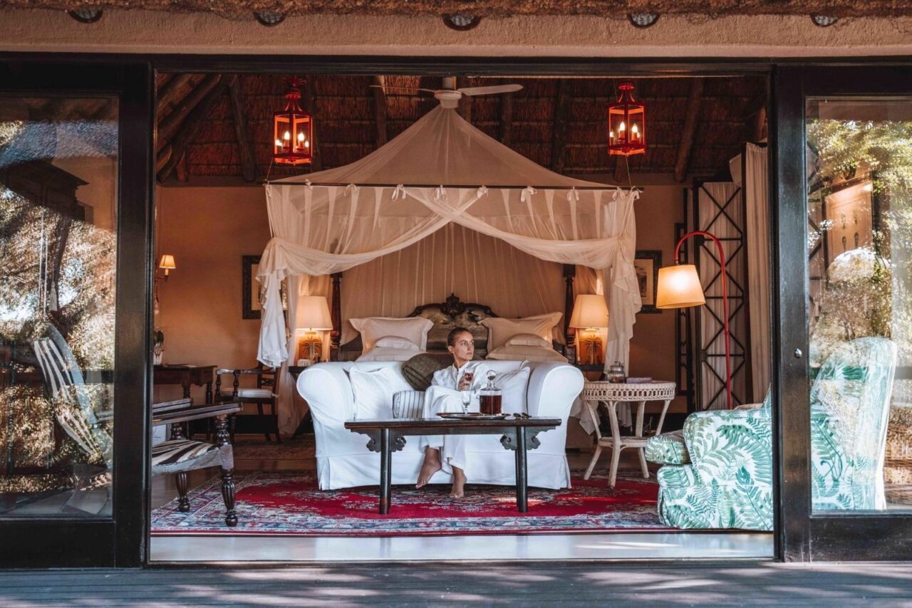 A woman sits on a white sofa in front of a white canopied bed. A red. rug is underneath her. This is one of the suites at Royal Malewane, one of the best safari lodges in South Africa. It's opulent with vintage decor and warm wood features.