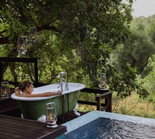 A woman sits in a green and white open air bathtub beside a swimming pool surrounded by trees.