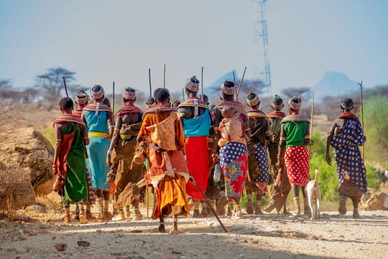 A group of Samburu women and children walk away from the camera along a dusty path, dressed in colorful traditional attire with intricate beadwork and patterned wraps. They carry sticks and calabashes, with one woman carrying a child on her back, against a dry landscape dotted with rocks and acacia trees.