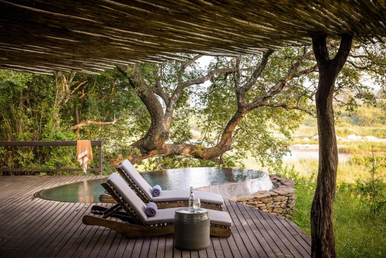 A pool at Singita Boulders. Two sunloungers sit in front of an oval pool with trees in the background.