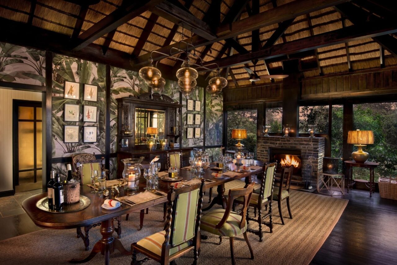 An opulent jungle themed dining room with dark wood ceiling and green leaves on the wallpaper. There is a long table in the centre of the room and a lit fireplace.
