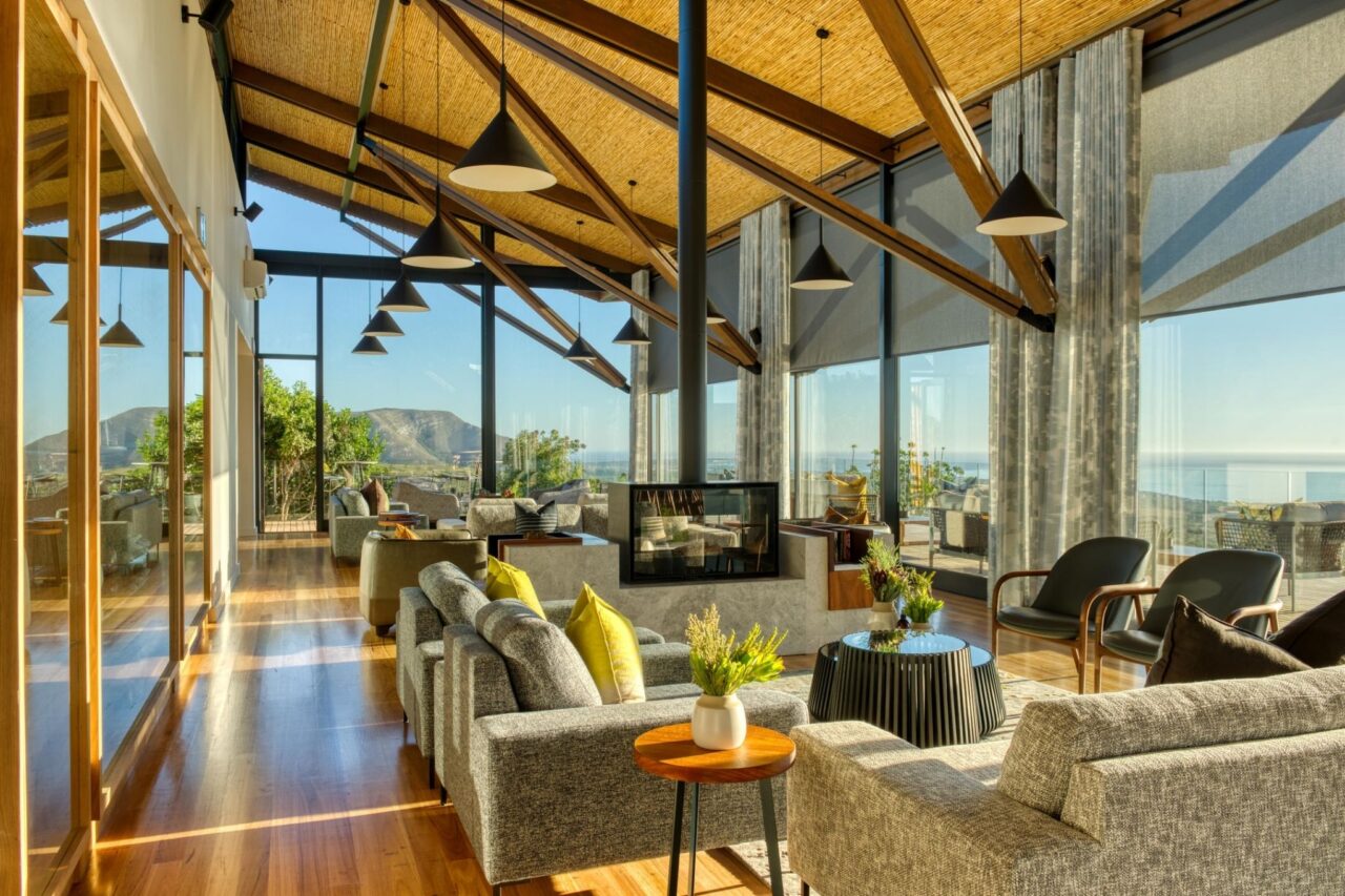 A large terrace/lounge area at The Garden Lodge with a high ceiling. Armchairs are set around a TV and the walls have floor to ceiling windows with views of the mountains.