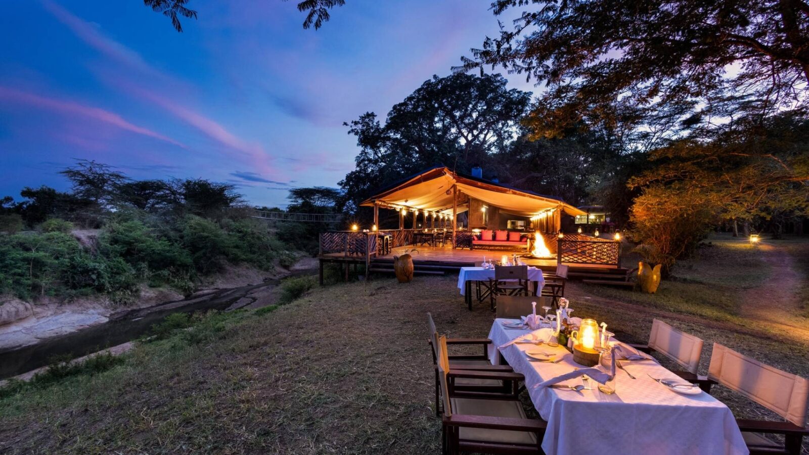 Saruni Basecamp Maasai Mara, Kenya. A table set outside for dinner lit up softly by lamps. A softly lit safari tent is behind it.