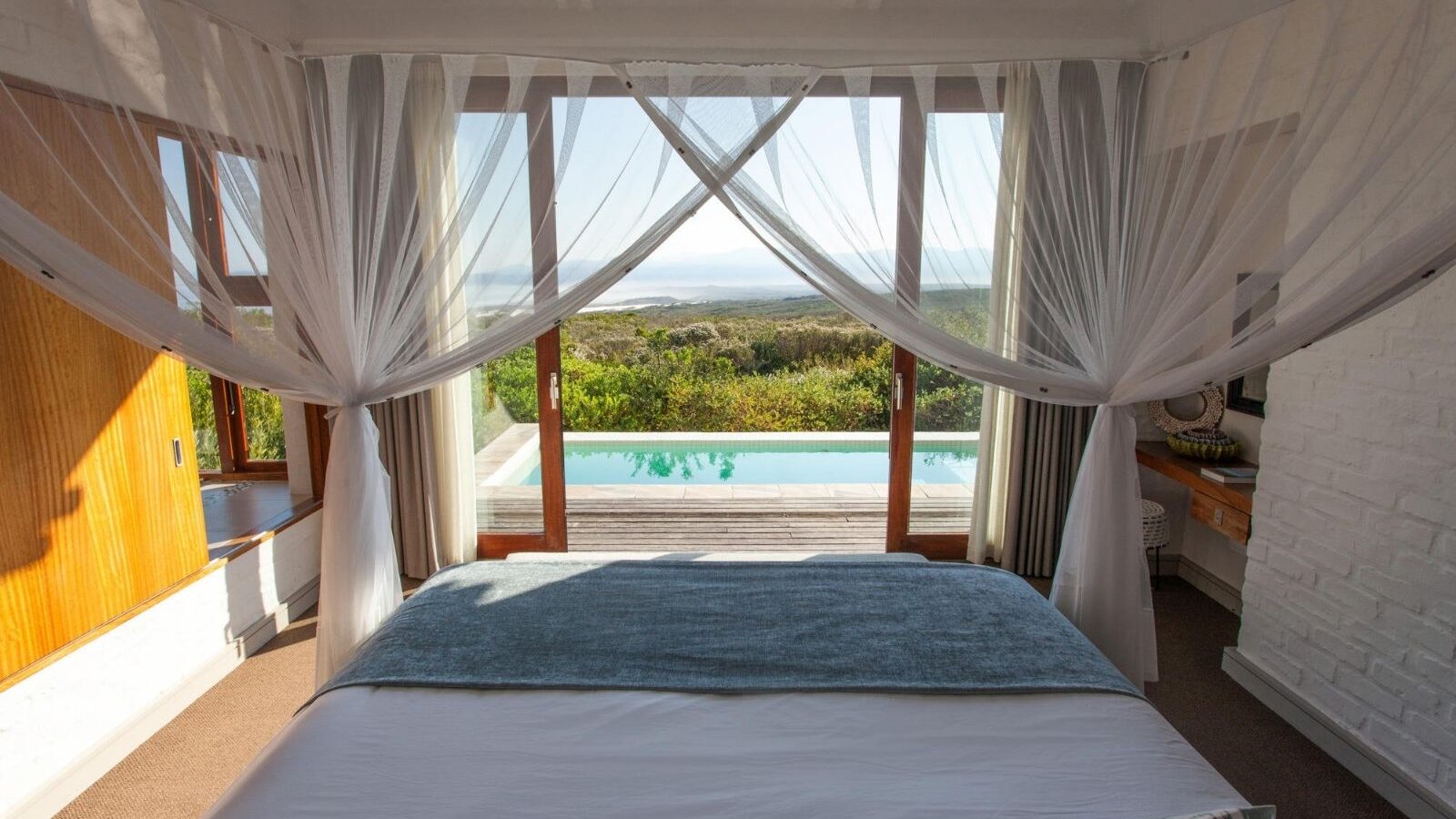 A bed looking out to a terrace with a pool on it and green forested hills beyond.