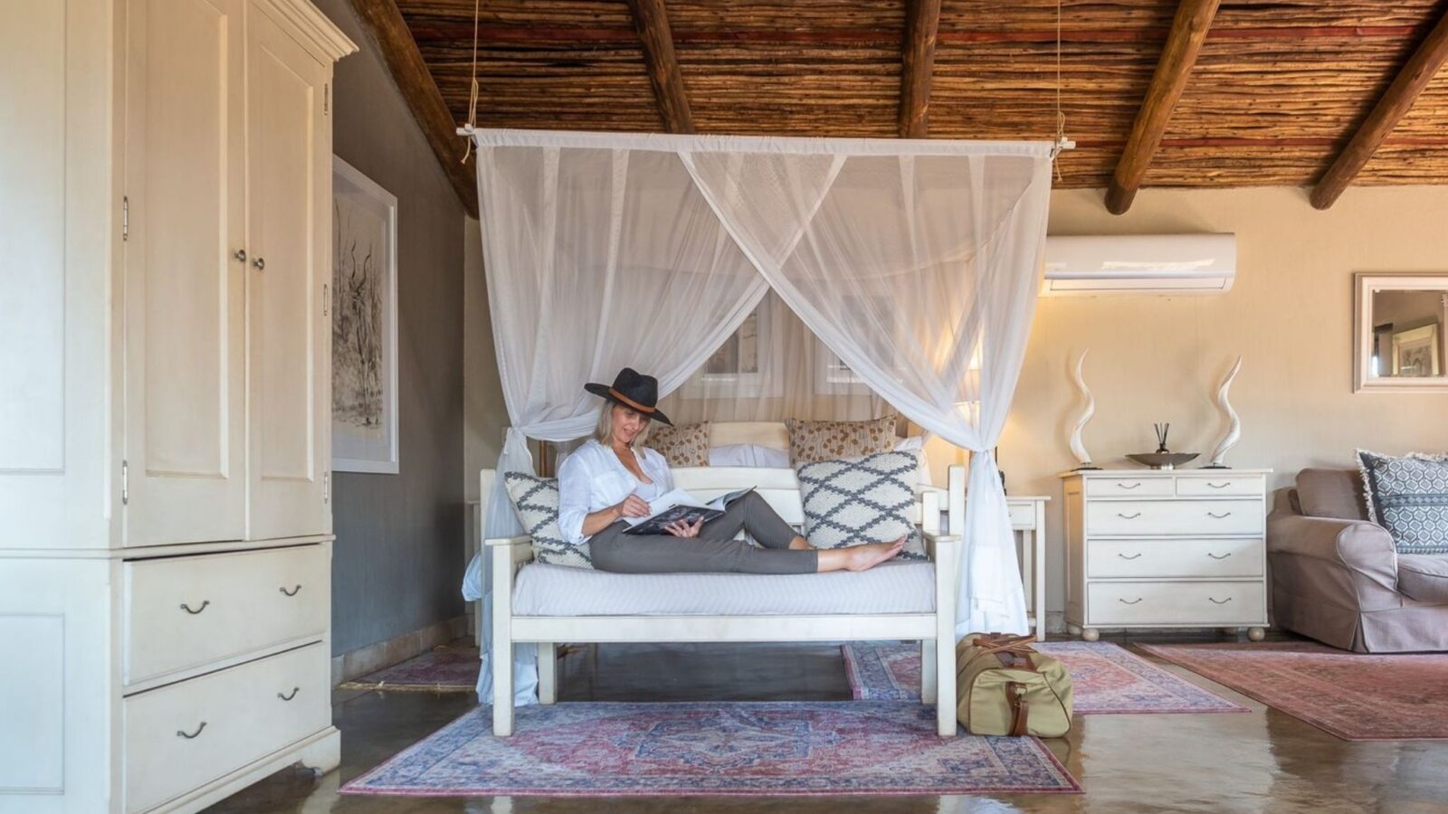 woman lounging on a sofa in front of a bed in a light room with wooden pannelled ceiling.