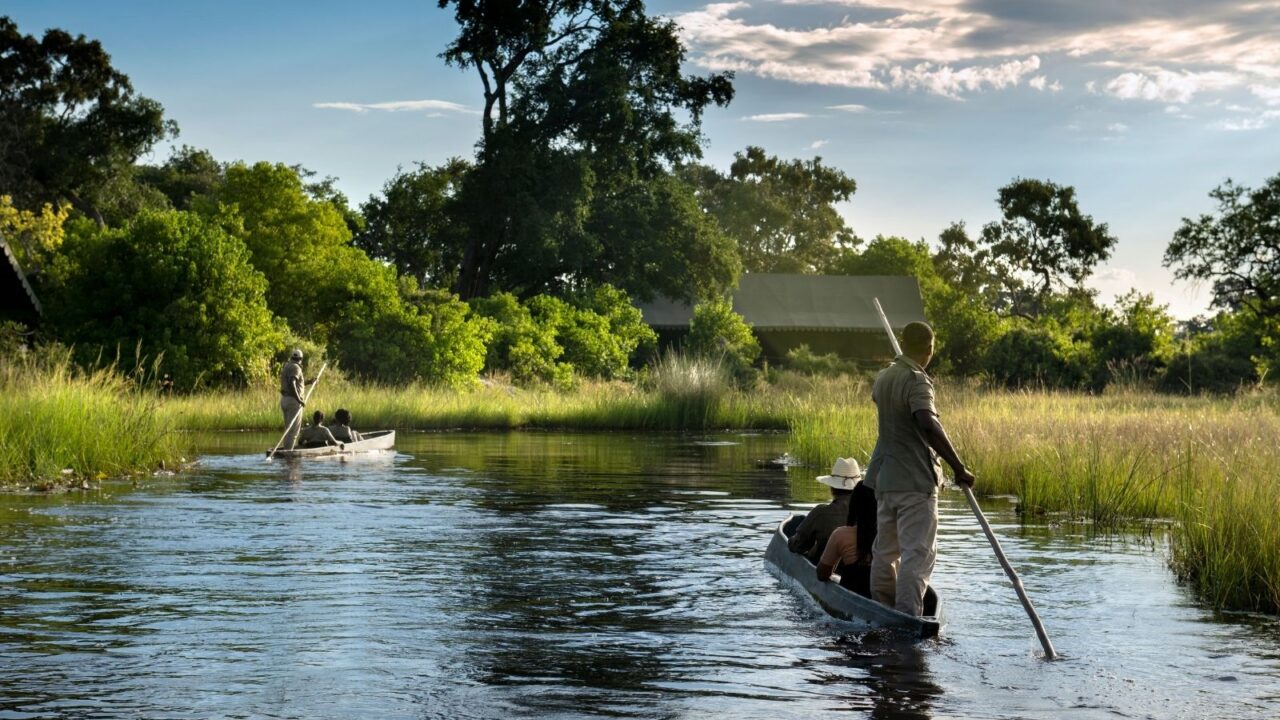 Jack's Camp. A guide stands in a mokoro canoe with a passenger sitting as the float on the Okavango Delta surrounded by the green bush.