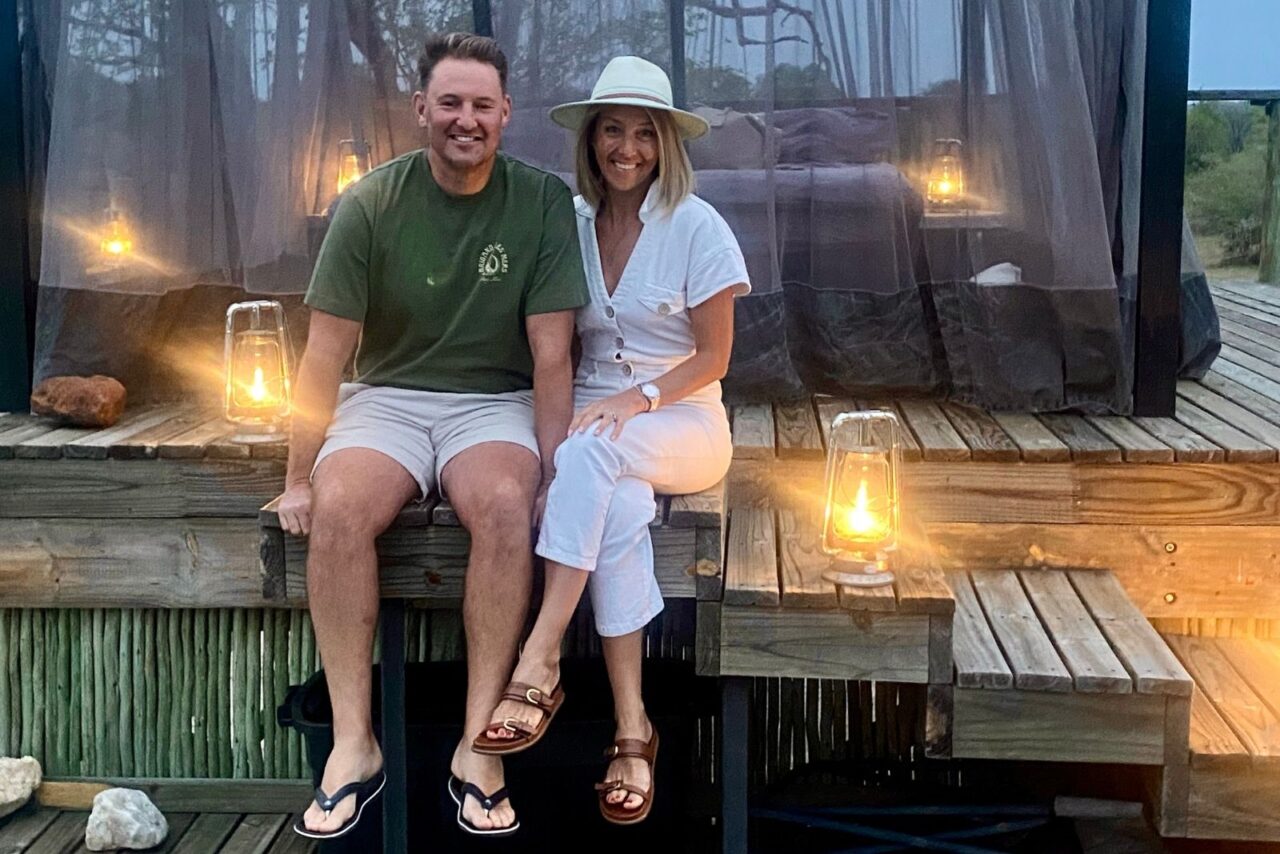 A couple sit surrounded by brightly lit lamps on their sleepout at Garonga. Fin wearing white shirts and a green t-shirt and Wendy wearing all white. Both are smiling happily.