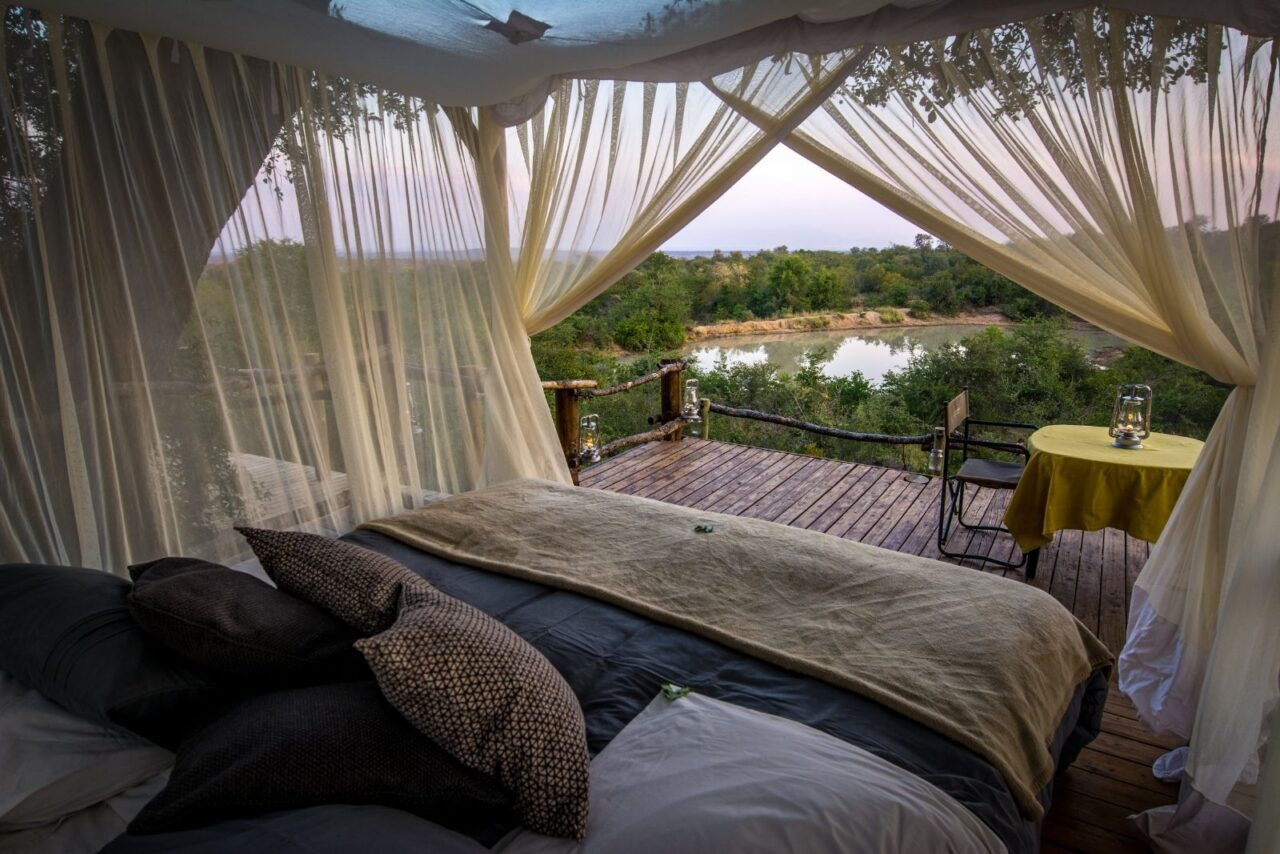 View from the sleepout bed at Garonga. It has a light canopy over the top and the deck is partially visible with the table. In the distance is a water hole surrounded by lush trees.